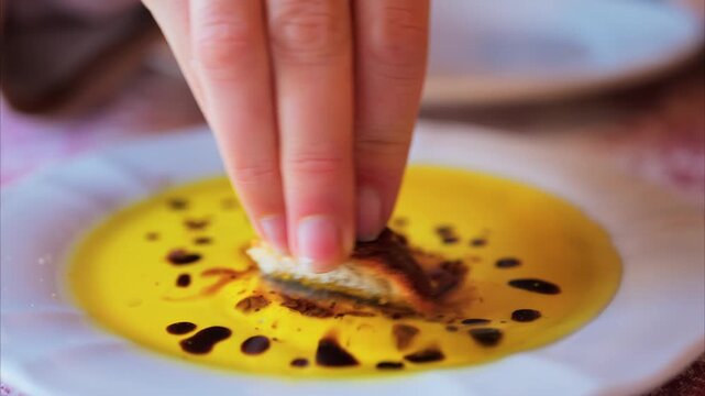 Person dipping fresh rustic bread into golden olive oil and balsamic vinegar on white plate.