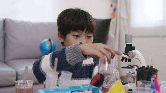 Happy adorable Asian boy with microscope sitting learning science and conducting chemistry experiments by mixing various colored liquids with test tubes and beakers, enjoying in nursery classroom lab.