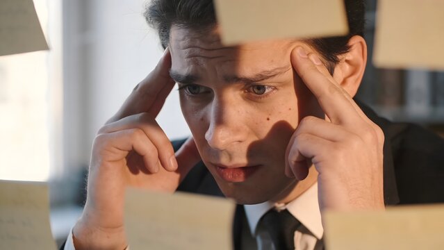 Stressed businessman looking at many sticky notes on a glass wall. Concept of burnout, information overload, deadline pressure, and project management challenges in a professional office setting
