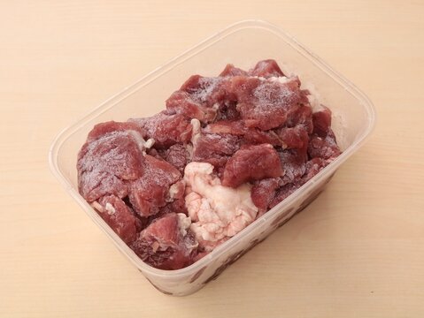 High angle view of fresh raw diced beef meat chunks with frost, stored inside a clear transparent plastic box container on a wooden table background, ready for cooking preparation
