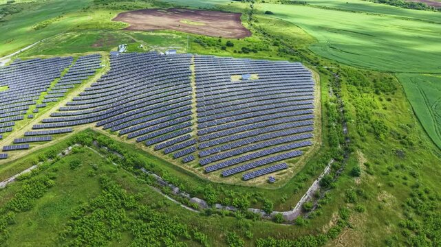 Utility solar array on grassy hill, dense rows of photovoltaic panels with service tracks and perimeter fence, close aerial perspective highlights industrial
