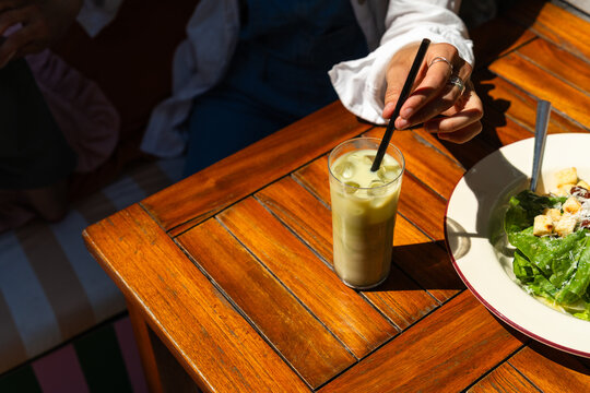 Person stirring iced matcha latte with a straw on a sunny restaurant terrace