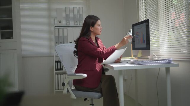 Focused Asian businesswoman sitting at her desk, comparing financial reports and analyzing data charts on a computer in a modern office