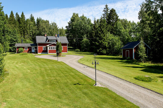 Traditional red wooden cottage with a gravel driveway, surrounded by green lawns and lush pine forests under a blue sky in Scandinavia. Typical Swedish summer countryside idyllic landscape.