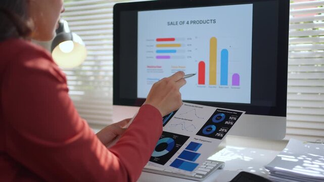 Young professional businesswoman comparing and reviewing financial statistics, charts, and graphs on a computer monitor and a paper document