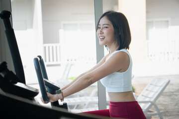 Fototapeta premium Happy young Asian woman in sportswear smiling and riding a stationary exercise bike in a gym, looking forward while enjoying her cardio workout routine for health and fitness