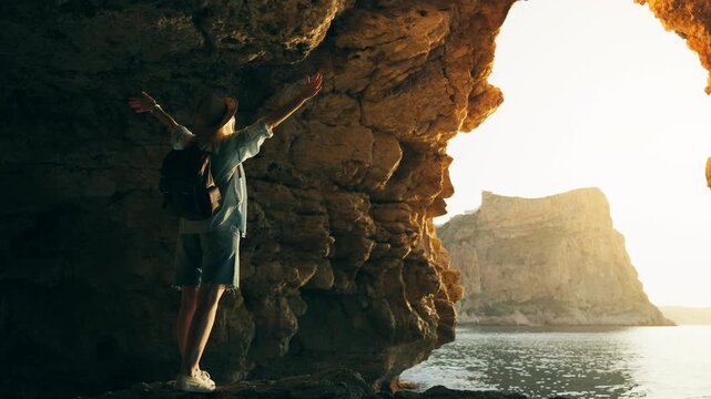 Young adventurous traveler woman with backpack exploring sea cave and raising arms in celebration while watch sunset over water, enjoy summer travel adventure, freedom, coastal vacation lifestyle.