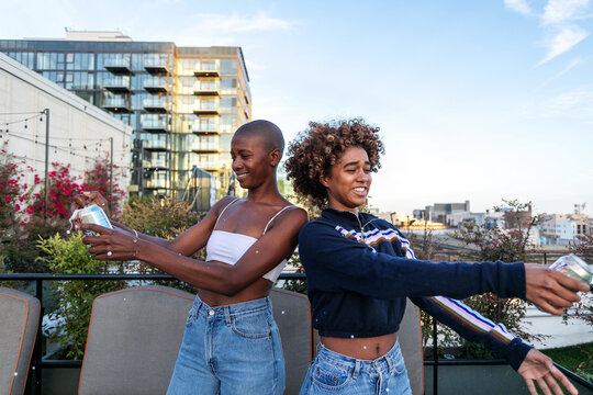 Two women smiling and holding drinks on a rooftop with city views.