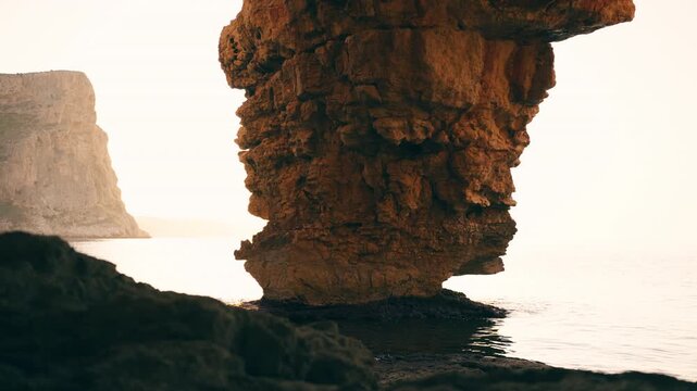 Stunning sea stack rock formation standing in the calm ocean water during a beautiful golden hour sunset, with imposing cliffs creating a serene and majestic coastal landscape