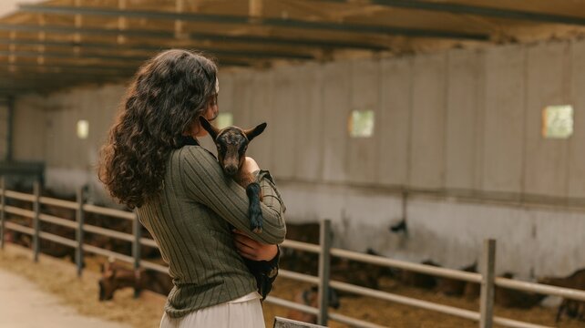 Young woman holding a baby goat in a barn with other animals in the background