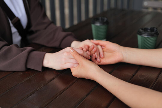 Two individuals sit opposite each other at a wooden table. They hold hands gently while coffee cups sit nearby. The scene takes place in a cafe during the day