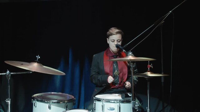 Energetic female drummer performing live on stage, singing while playing the drum kit under dramatic concert lighting.