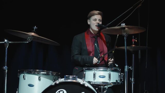 Energetic female drummer performing live on stage, singing while playing the drum kit under dramatic concert lighting.