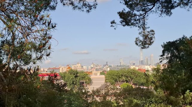 Panoramic look at Beijing cityscape from Jingshan hill through fresh tree leaves. Famous Chinese capital metropolis overview under clear sky, showcasing contrast of historic park and downtown