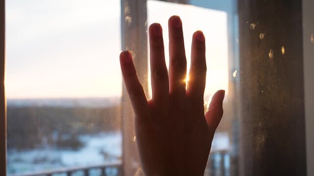 Backlit silhouette of persons hand touching window with warm sunset shining through fingers. Creating lens flares while looking out at a snowy winter landscape. Gesture of helping hand and hope