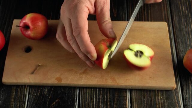 Close-up of hands slicing a red apple in half on a wooden cutting board with additional apples surrounding the scene in a rustic kitchen setting