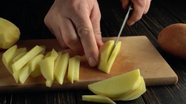 Hand slicing fresh potatoes into thin strips on a wooden cutting board with a rustic kitchen background and additional potatoes visible
