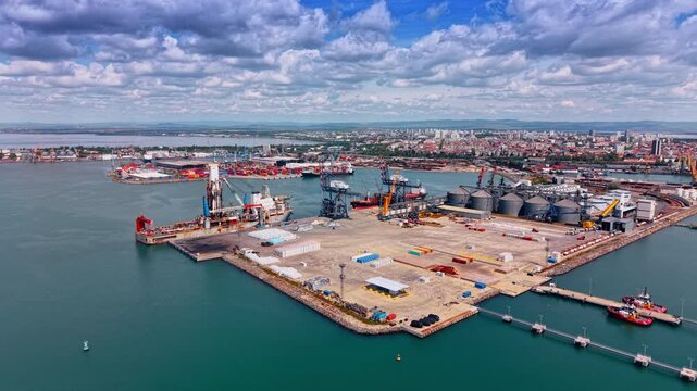Flying away from large open area for containers in the modern port. Cranes and huge metal tanks around. Port and cityscape under the cloudy sky.