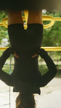 A person hangs upside down from yellow monkey bars in an outdoor park setting. They are engaged in playful exercise or a workout at the playground