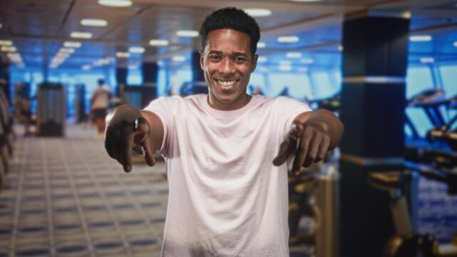 Man pointing both index fingers at camera in gym, smiling, visible forearms and casual pink t shirt, cardio machines behind him; confidence invitation.