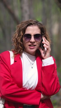 A charming curly young woman, a businesswoman in a red jacket, talks on the phone against the backdrop of the greenery of a summer park.