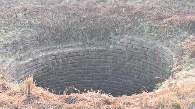 An old deep brick built water well. Its circular interior wall is lined with weathered, dark bricks, while the surrounding surface is covered in dry grass, straw, and wild vegetation.