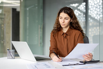 A young businesswoman sits at a desk with a laptop, papers, and a clipboard, writing notes while...