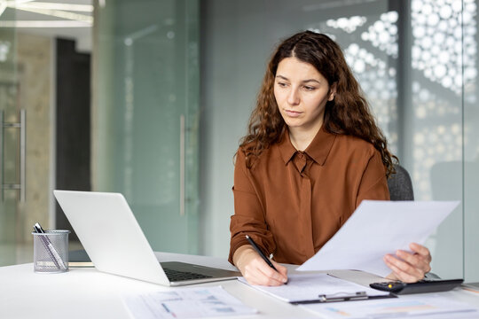 A young businesswoman sits at a desk with a laptop, papers, and a clipboard, writing notes while reviewing documents in a modern office environment