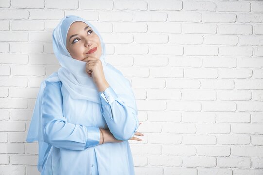 Muslim woman in blue hijab thinking and looking up against a white brick wall background