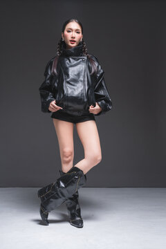 Full length of confident Asian woman in leather jacket and boots for luxury outerwear marketing campaign strong stance fashion aesthetic modern urban lifestyle bold portrait studio