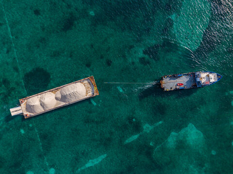 Aerial view of a tugboat towing a barge loaded with white sand across the clear turquoise waters near Arawak Cay in Nassau, New Providence, The Bahamas.