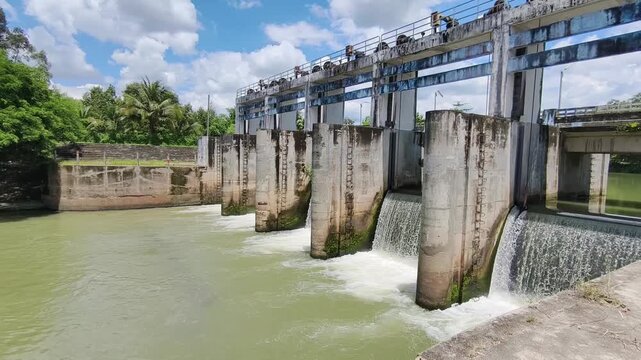 A large concrete dam with water flowing through multiple sluice gates under a bright blue sky with some clouds