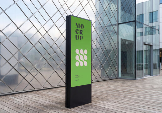 Perspective view of a vertical black outdoor digital signage totem mockup standing on a wooden deck in front of a modern glass building facade