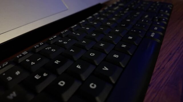 Teclado de laptop con iluminaci&oacute;n azul sobre escritorio de madera en ambiente oscuro, vista en perspectiva