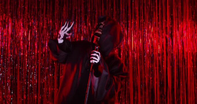 Grim reaper in black cloak with skull death mask dancing at Halloween party. Skeleton spooky monster in black cloak playing scythe like guitar of rock musician in front of red tinsel curtains
