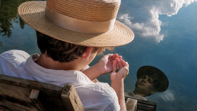 Adolescent wearing boater hat gazing at reflection in serene lake, clutching a small frog in his hands