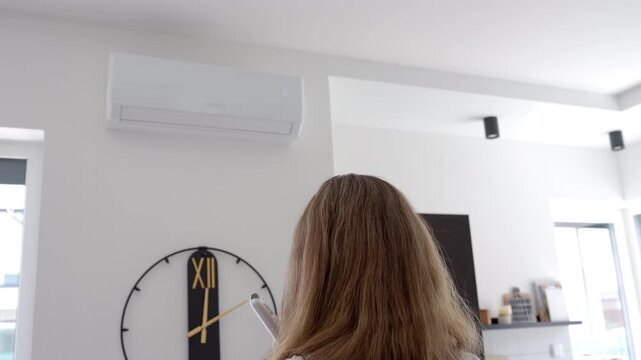 Woman using remote control to adjust temperature on wall mounted air conditioner in bright room. Concept of cooling, summer heat and temperature control