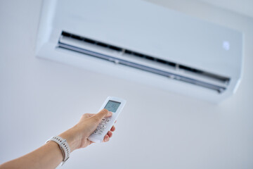 Hand adjusts a wall air conditioner with a remote control in a bright room. Concept of cooling,...