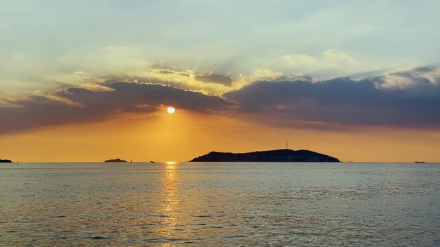 Kinaliada at sunset beneath dramatic clouds, golden reflections and radiant sunbeams creating atmospheric timelapse seascape. Cargo ship silhouette crossing calm sea 