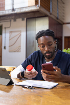 African American man in scrubs holding pill bottle sitting at table in loft checking red phone