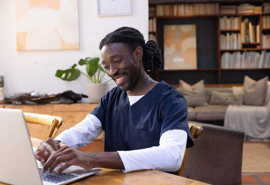 African American man sitting at wooden table using silver laptop in living room wearing navy scrubs
