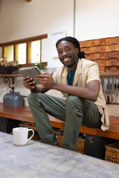 Adult African American man holding tablet and smiling toward camera on wooden bench near mug