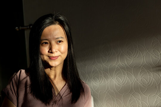 Mature Asian woman smiling in lit room, mauve top, textured wall, sunlight shadows, copy space