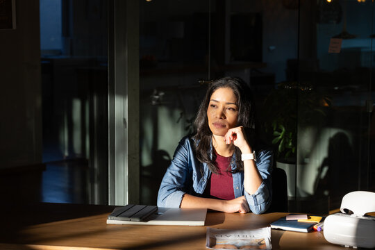 African American woman wearing denim jacket sitting at coworking desk with closed laptop, pondering