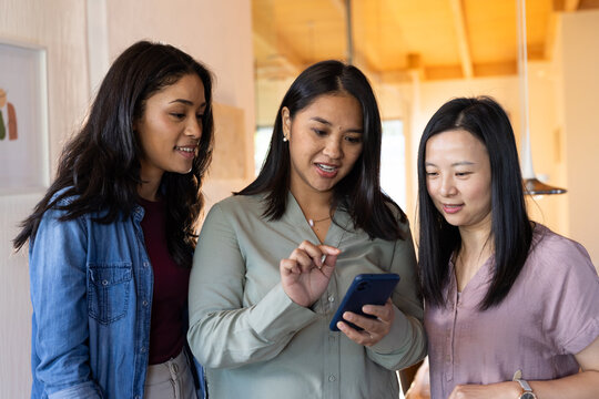 Asian women coworkers leaning in while tapping blue smartphone at warm office with pendant light