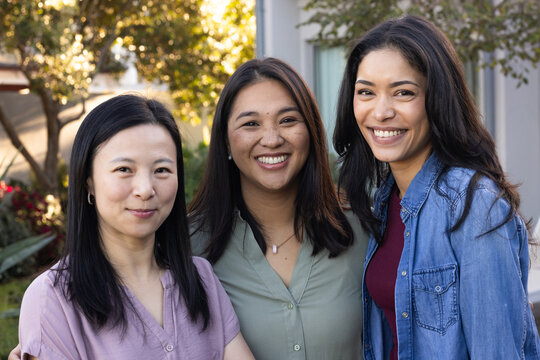Diverse female friends standing close together in yard wearing mauve blouse, pendant, denim jacket