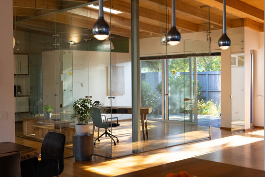 Glass-walled office is featuring wooden desk and rolling chair in sunlit workspace, with plants
