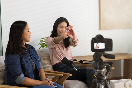 Two African American women presenting bottle toward DSLR camera on sofa, microphone visible