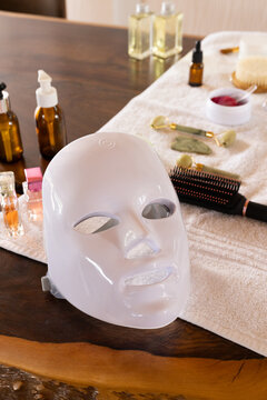 White glossy LED facial mask is resting on live-edge table with amber bottles and gold roller