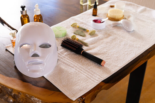 White LED face mask is resting on towel on live-edge table with paddle brush, jade roller